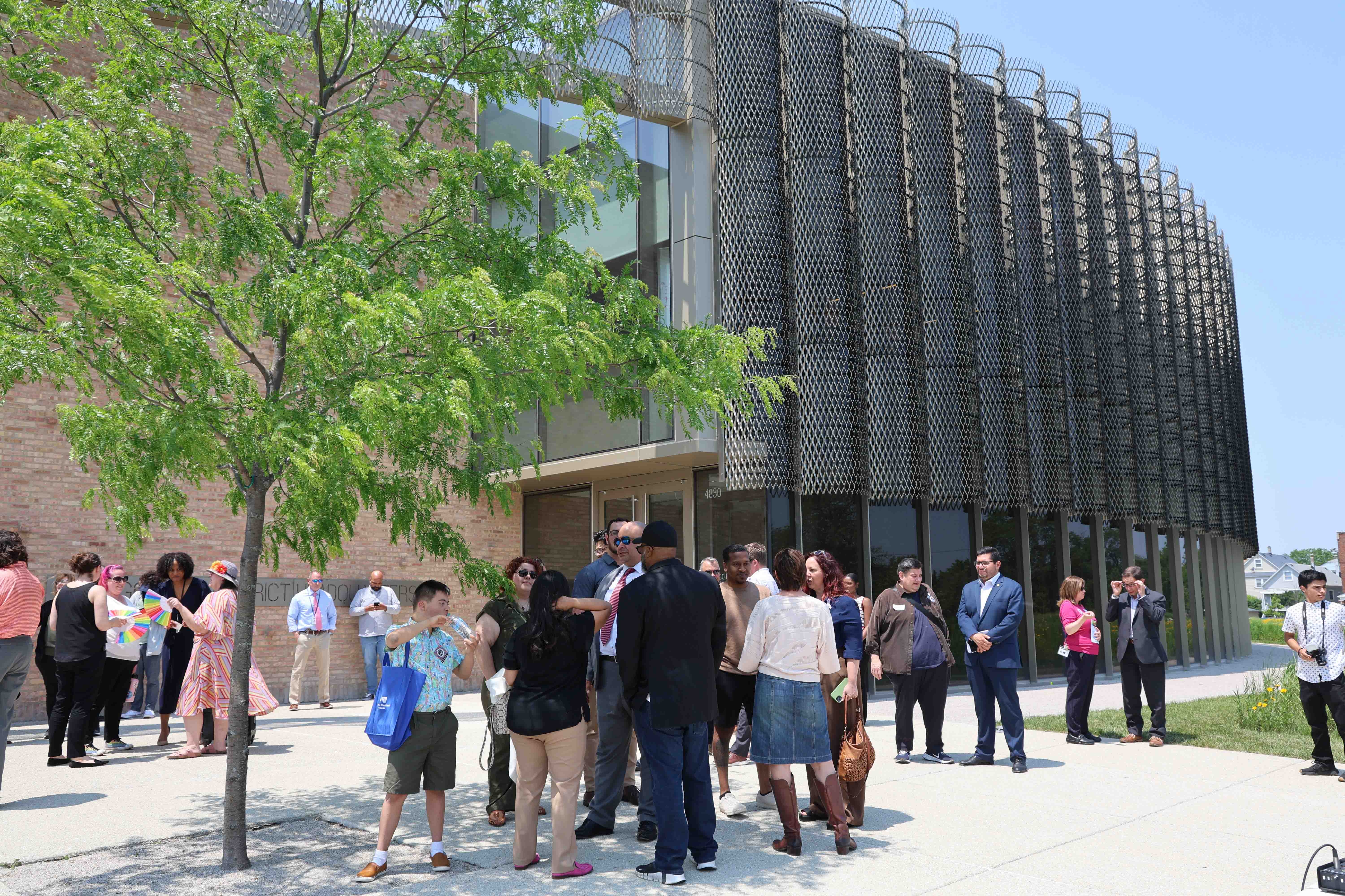People gather outside a modern building with a curved metal screen facade.